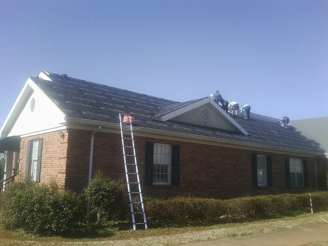 Crew tearing shingles off a brick ranch roof with a ladder set — Emergency Roof Repair work by our crew in Newman Grove