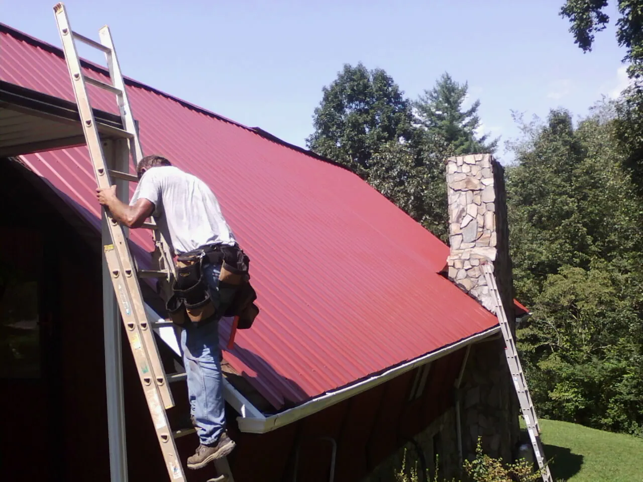 Roofer on a ladder beside a red metal roof and stone chimney — Soffit & Fascia Repair by Roofing Brothers in Newman Grove